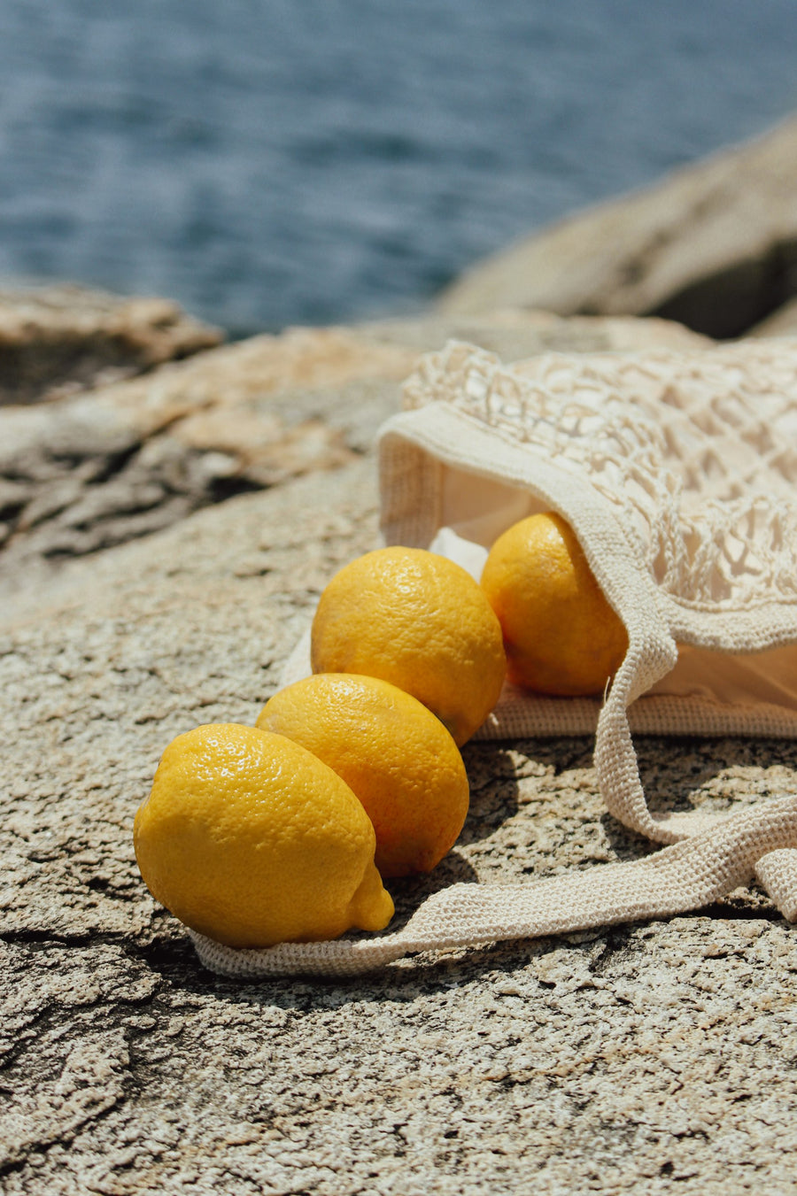 Fresh lemons spilling from a cotton net bag onto sunlit sea rocks, for Luna & Lo. fresh and citrus candles