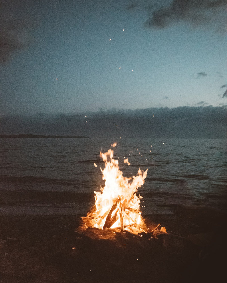 Driftwood bonfire glowing on the shoreline at dusk, for Luna & Lo. woody and smoky candles